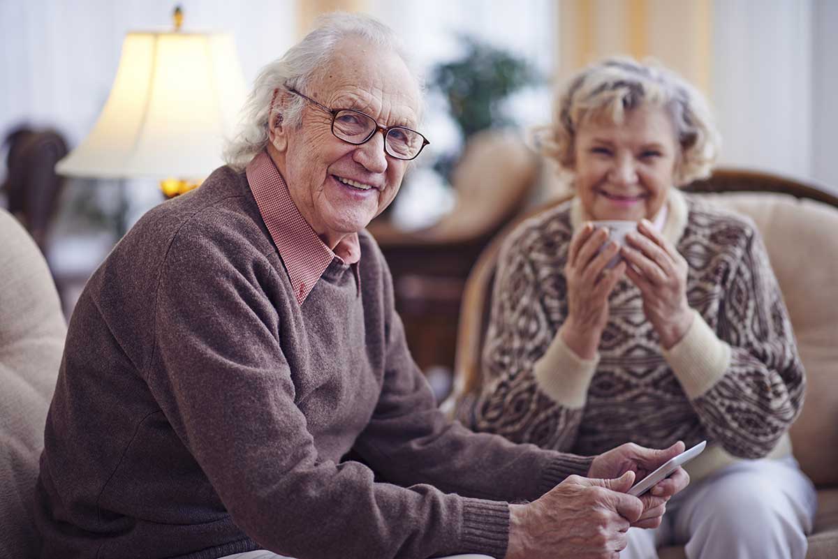 senior man with touchpad looking at camera with his wife sitting in background