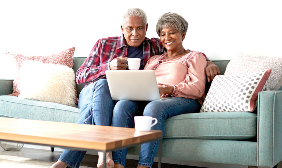 Senior Couple Sitting On Sofa At Home Using Laptop To Watch Senior Choice at Home Online Event