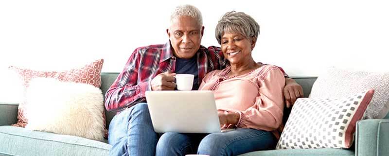 Senior Couple Sitting On Sofa At Home Using Laptop To Watch Senior Choice at Home Online Event