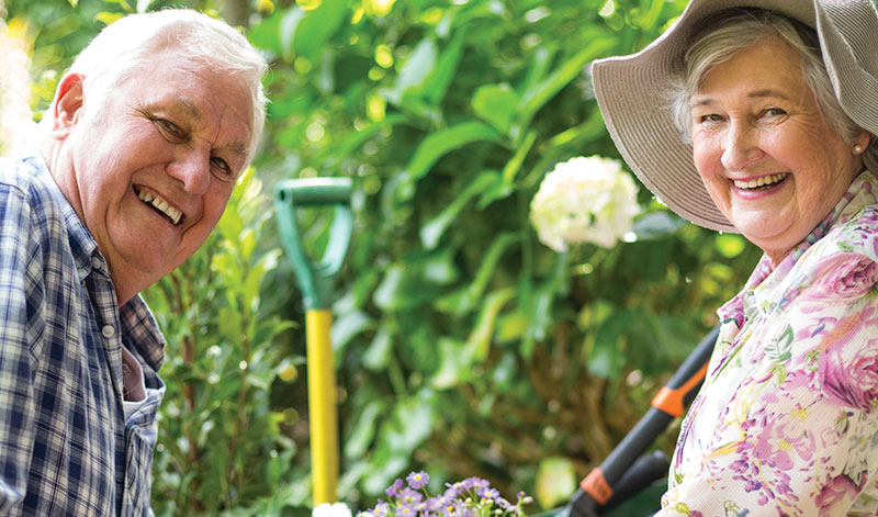 An elderly man and woman are smiling at the camera while gardening. They are surrounded by greenery and colorful flowers. The woman is wearing a wide-brimmed hat.