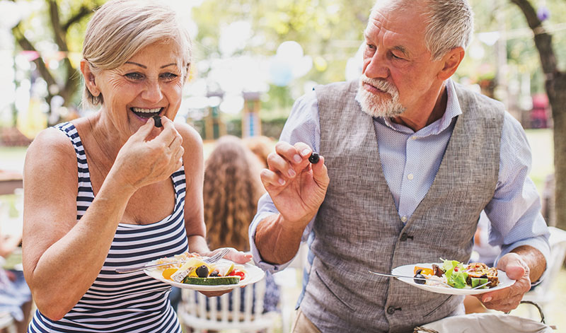 An elderly couple enjoys a meal outdoors, with the woman eating a bite of food and the man holding a piece of food. Other people are blurred in the background.