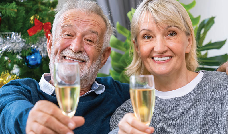 An older man and woman smile while holding glasses of champagne. A decorated Christmas tree is visible in the background.