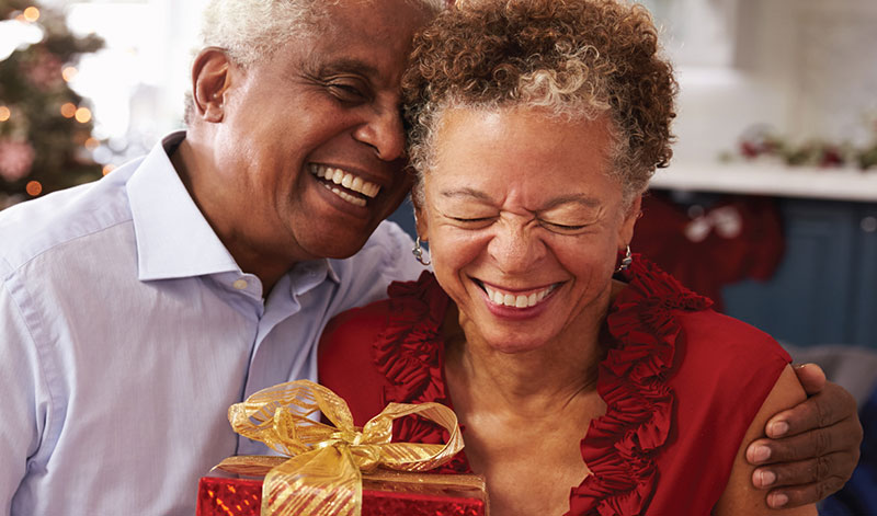 An elderly couple shares a joyful moment. The man hugs the woman, who is smiling with her eyes closed and holding a gift wrapped in red paper with a golden ribbon.