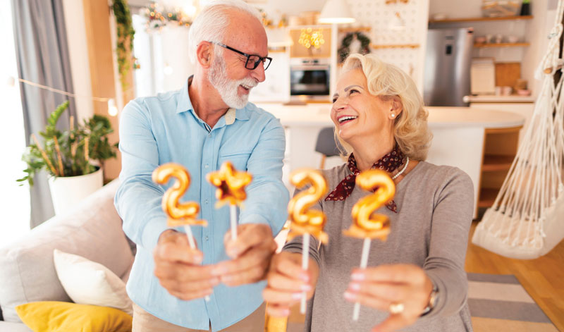 An elderly couple stands in a kitchen, holding golden balloon numbers "2," "0," "2," and "2," while smiling at each other.