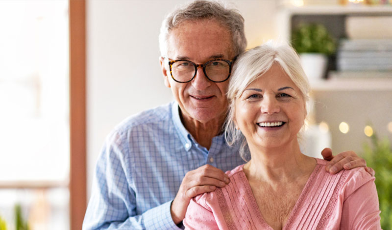 An elderly couple, the man wearing glasses and a blue shirt, and the woman in a pink top, smile warmly at the camera. They are indoors with a blurred background.
