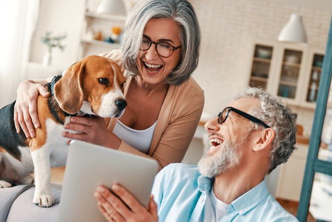 Two smiling older adults sitting indoors with a beagle, sharing a moment together while looking at a tablet.