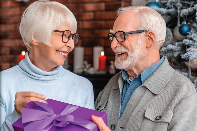 An elderly couple smiles warmly at each other as the woman holds a purple gift box with a ribbon. They stand in front of a decorated Christmas tree and lit candles.