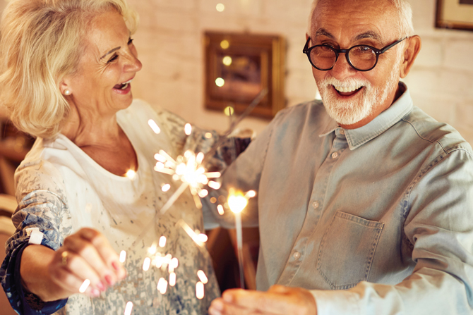An elderly couple smiles while holding sparklers indoors.