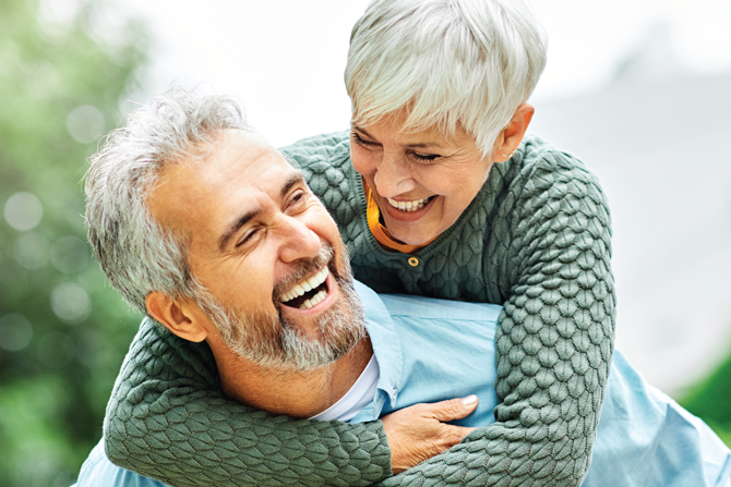 A gray-haired woman with a broad smile playfully embraces a laughing gray-haired man from behind while both wear casual sweaters outdoors.
