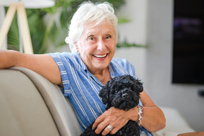 An elderly woman with white hair and a blue-striped shirt sits on a couch smiling, holding a small black dog.