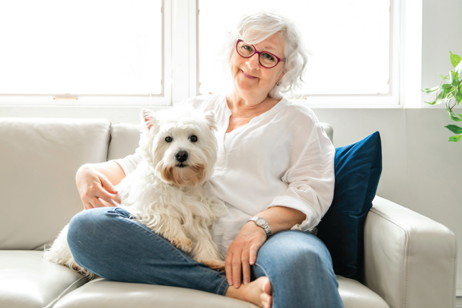 An elderly woman with short white hair and glasses sits on a sofa, holding a small white dog on her lap. Both are looking at the camera.